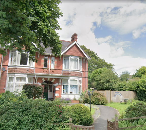 Red-brick exterior of Bryn Irfon with bay windows, a tiled gable and a front garden with shrubs and a path