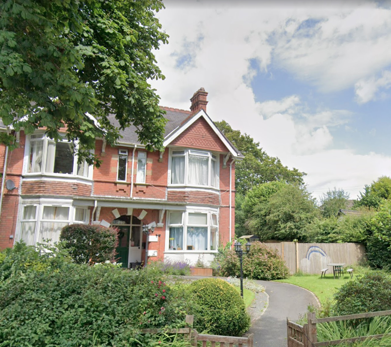 Red-brick exterior of Bryn Irfon with bay windows, a tiled gable and a front garden with shrubs and a path