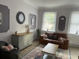 Sitting room at Brynawel with a tan leather sofa, grey armchair, sideboard and a large wall clock