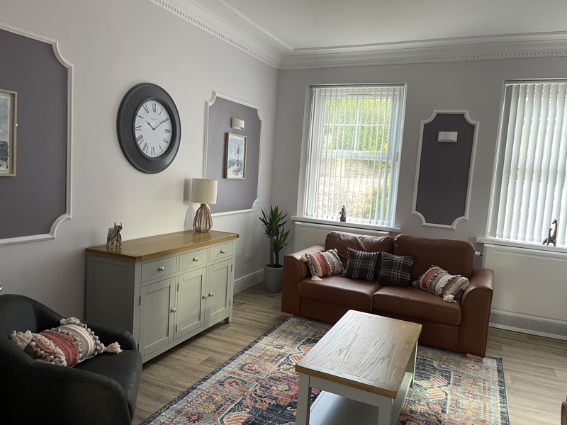 Sitting room at Brynawel with a tan leather sofa, grey armchair, sideboard and a large wall clock