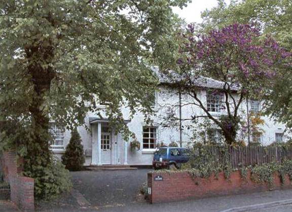 Exterior of The Grove, a white-rendered house behind mature trees with a low brick wall and a car parked outside
