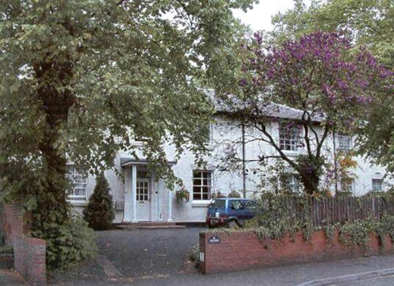 Exterior of The Grove, a white-rendered house behind mature trees with a low brick wall and a car parked outside