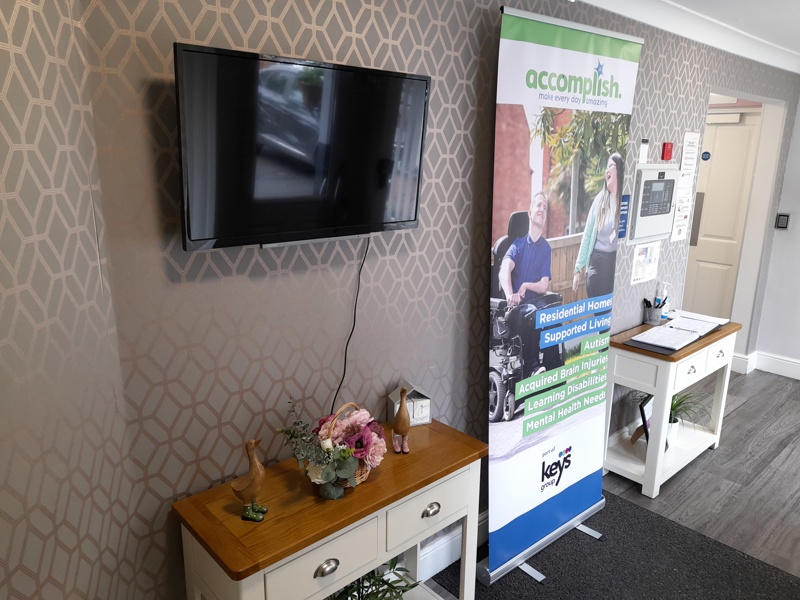 Reception lounge at Boston House with a wall-mounted TV, console table and an Accomplish information banner