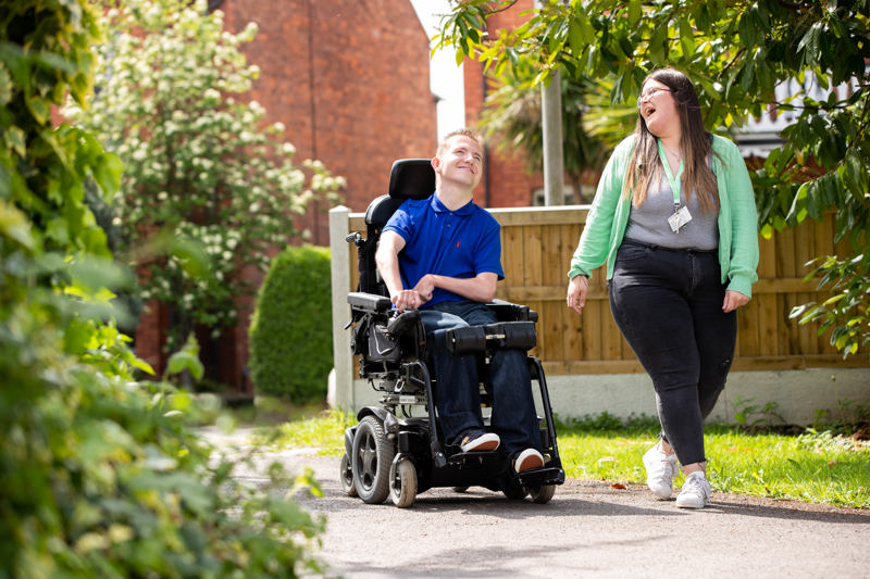 A man in a powered wheelchair and a support worker walking along a garden path beside the building at Sandpiper