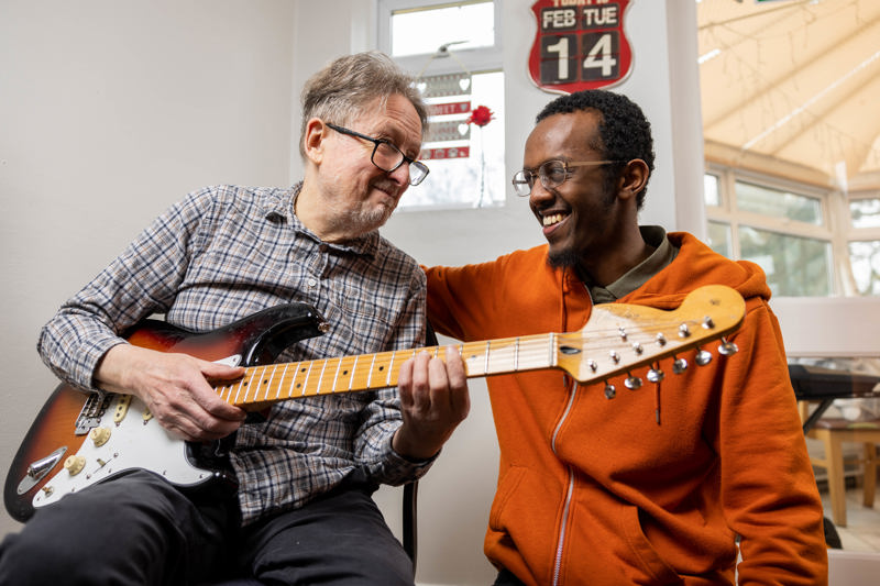 Two men playing electric guitars together in a sunlit lounge at Cae Deri