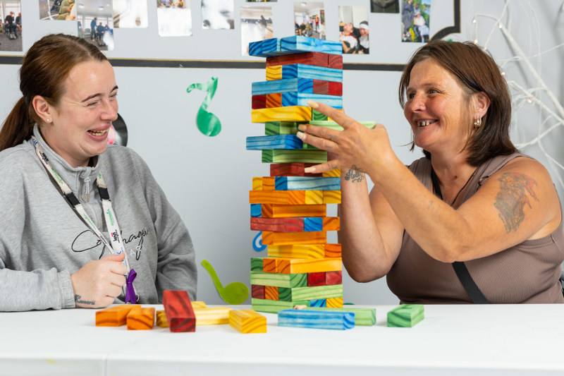 Two women laughing while playing a giant Jenga tower of rainbow blocks at a table at Sandpiper