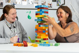 Two women laughing while playing a giant Jenga tower of rainbow blocks at a table at Paddocks