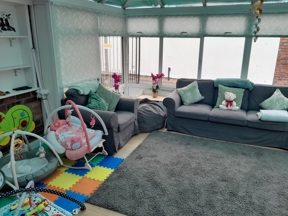 Bright conservatory living room at Victoria Villas with grey sofas, a baby bouncer and colourful play mats