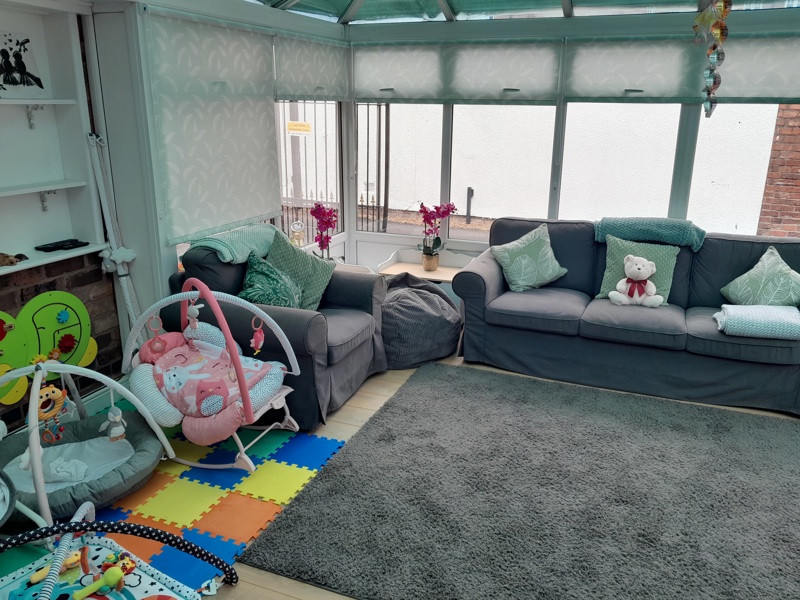 Bright conservatory living room at Victoria Villas with grey sofas, a baby bouncer and colourful play mats