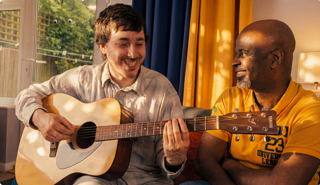 Two people sitting indoors smiling while one plays an acoustic guitar. Blue and yellow curtains and a window with greenery visible behind them.