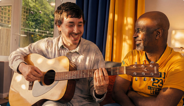 Two people sitting indoors smiling while one plays an acoustic guitar. Blue and yellow curtains and a window with greenery visible behind them.