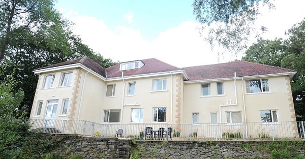 Exterior of Taith Cartref, a large cream-rendered care home with a tiled roof, patio seating and surrounding trees