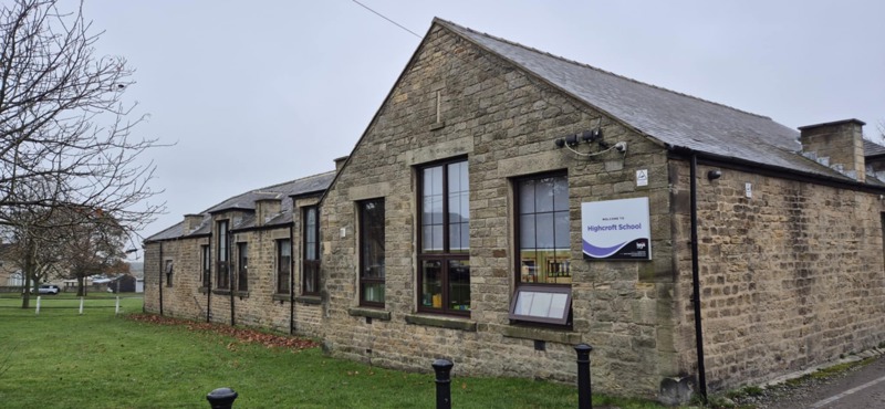 Stone-built single-storey Highcroft School with a welcome sign, lawn and bare tree under a grey sky