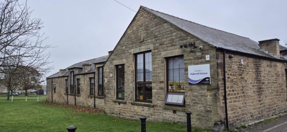 Stone-built single-storey Highcroft School with a welcome sign, lawn and bare tree under a grey sky