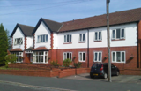 Front exterior of Byron Lodge, a white and red-brick two-storey care home with on-street parking