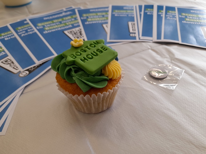 Celebration cupcake topped with a green Boston House fondant plaque, beside promotional leaflets on a table
