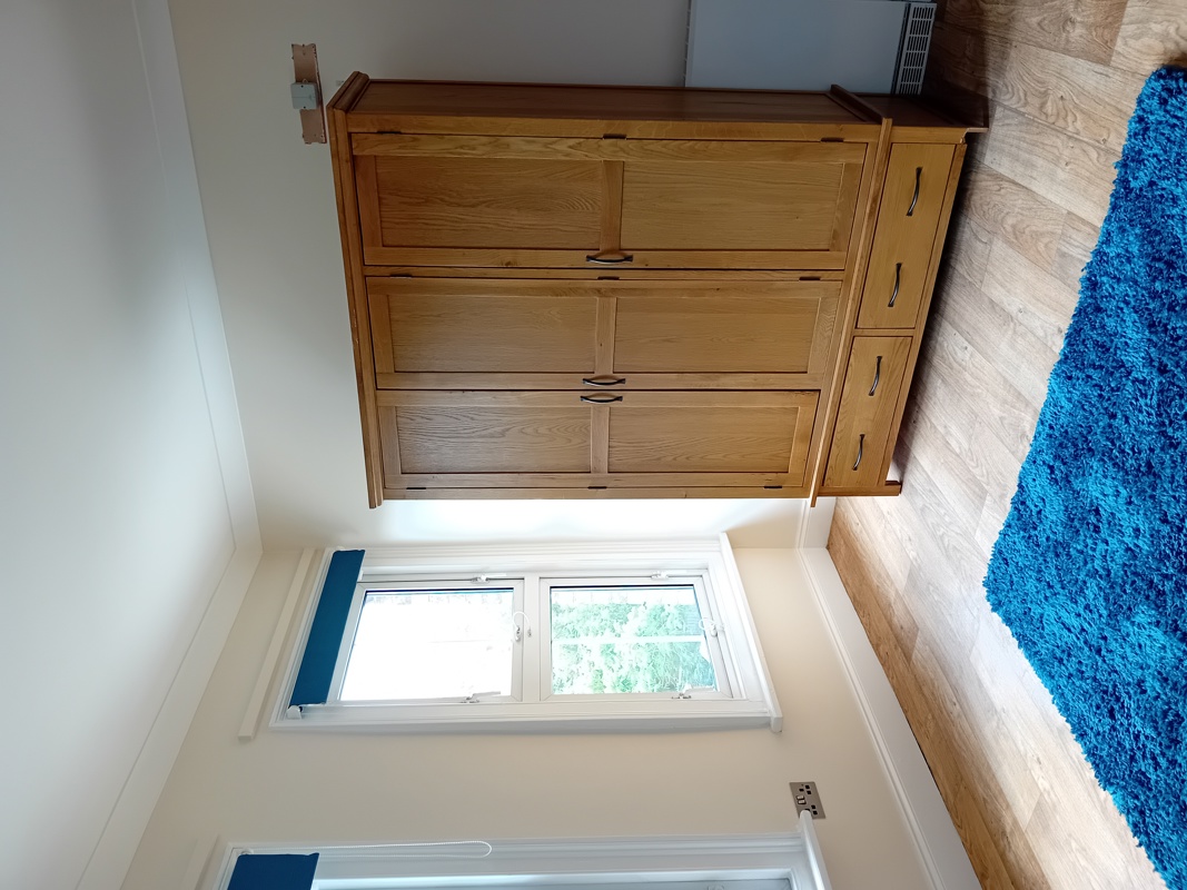 Bedroom at Eynesbury House with a large oak chest of drawers, a window and a bright blue rug