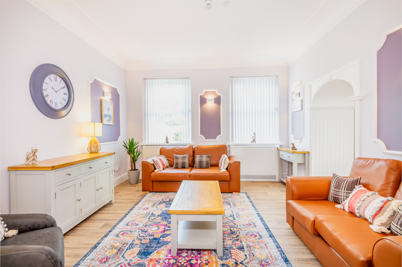 Lounge at Brynawel with tan leather sofas, a patterned rug, a large wall clock and lilac wall panels
