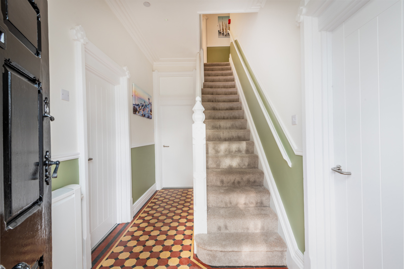 Entrance hallway at Brynawel with a Victorian tiled floor, carpeted staircase and a sage green dado