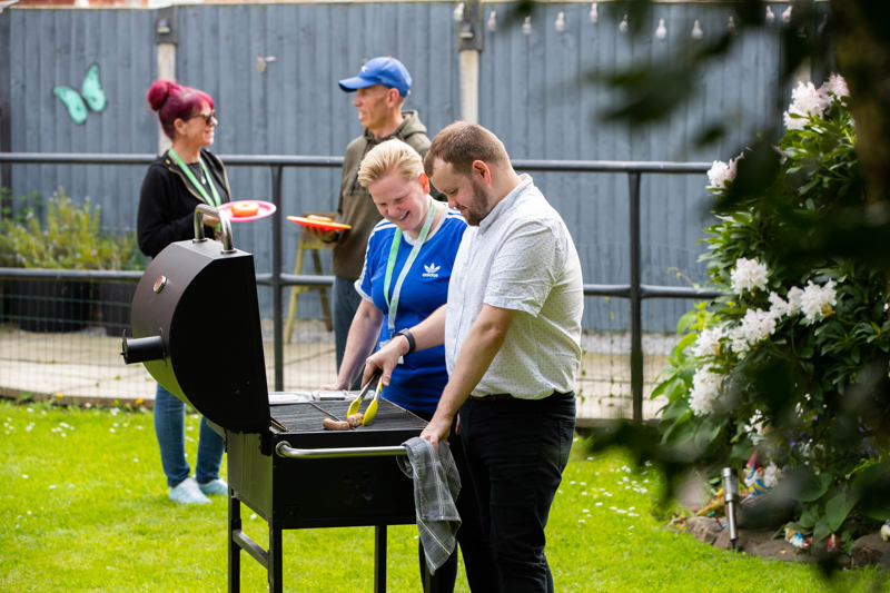Residents and staff at a garden barbecue at Orchard Hill, grilling sausages on a lawn beside flowering shrubs