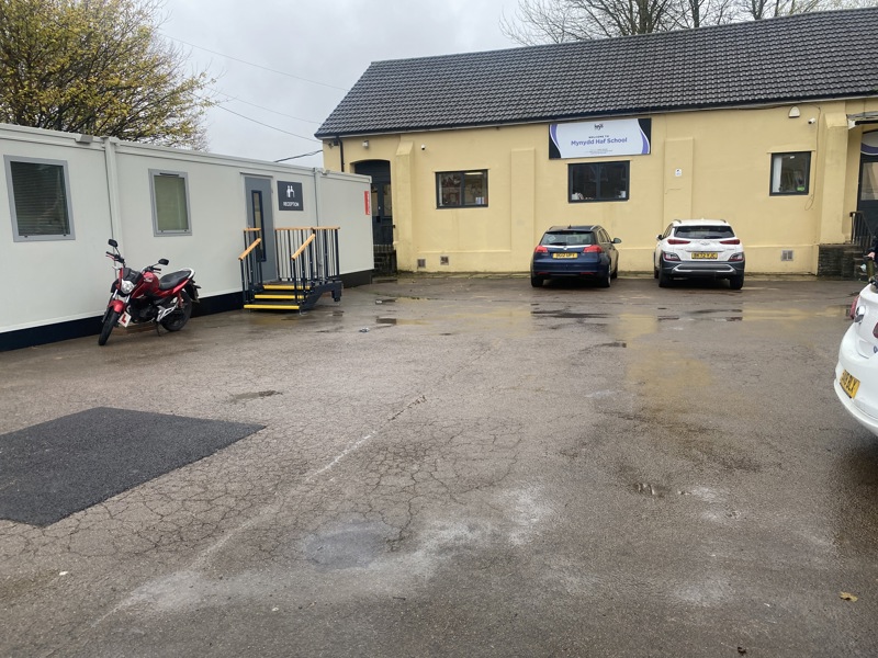 Cream-rendered Mynydd Haf School building with a welcome sign, reception portacabin and cars in the car park