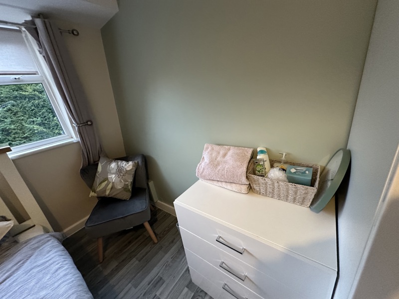 Bedroom at Evergreen with a white chest of drawers, toiletries basket and a grey armchair by the window