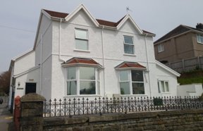 Front exterior of Gwynfryn, a white rendered house with bay windows and ornate iron railings on a stone wall