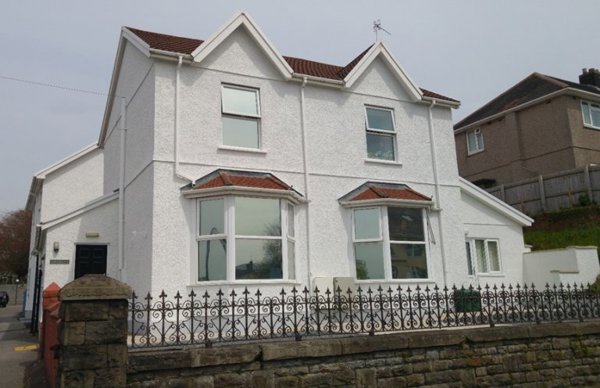 Front exterior of Gwynfryn, a white rendered house with bay windows and ornate iron railings on a stone wall