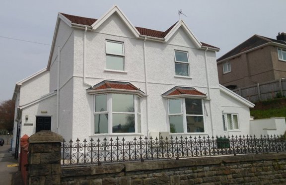 Front exterior of Gwynfryn, a white rendered house with bay windows and ornate iron railings on a stone wall