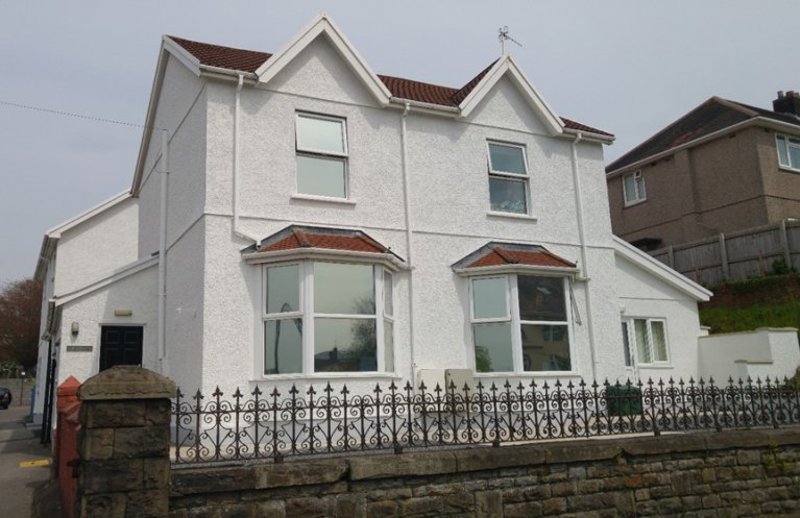 Front exterior of Gwynfryn, a white rendered house with bay windows and ornate iron railings on a stone wall