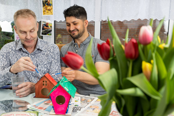 Two men painting wooden birdhouses at a craft table with tulips in a South West supported living conservatory