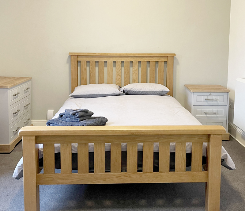 Light coloured wooden bed with grey duvet and 2 bedside drawers
