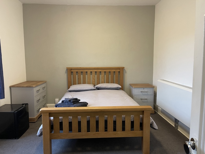 Bedroom at Glanmore with a double wooden bed, chest of drawers, bedside table and mini fridge