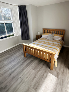 Light coloured wooden bed with striped orange duvet on a grey floor with window looking onto a garden