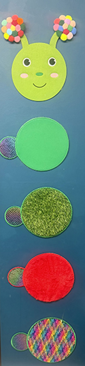 Textured sensory caterpillar craft with coloured fabric circles on a blue display board at Pinetrees