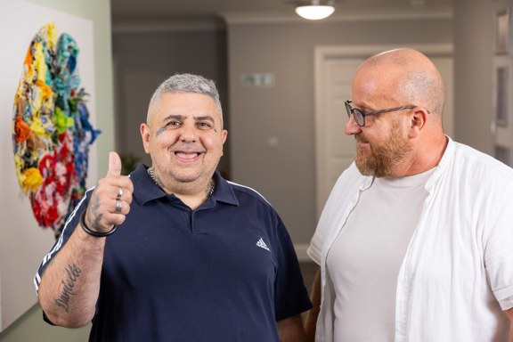 Two men in a bright hallway at Paddocks, one giving a thumbs up beside colourful textile wall art