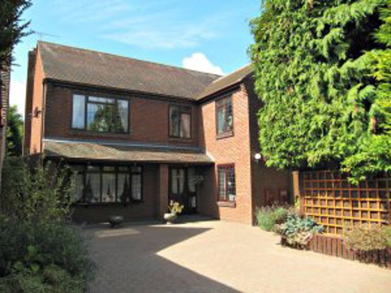 Exterior of The Orchard, a red-brick detached house with a bay window, paved driveway and conifers
