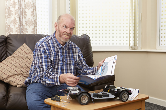 Man building a model car kit with instructions on a sofa in a Merseyside supported living lounge