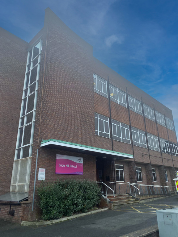 Brown-brick three-storey exterior and entrance of Snow Hill School with a pink sign and a tall glazed stairwell