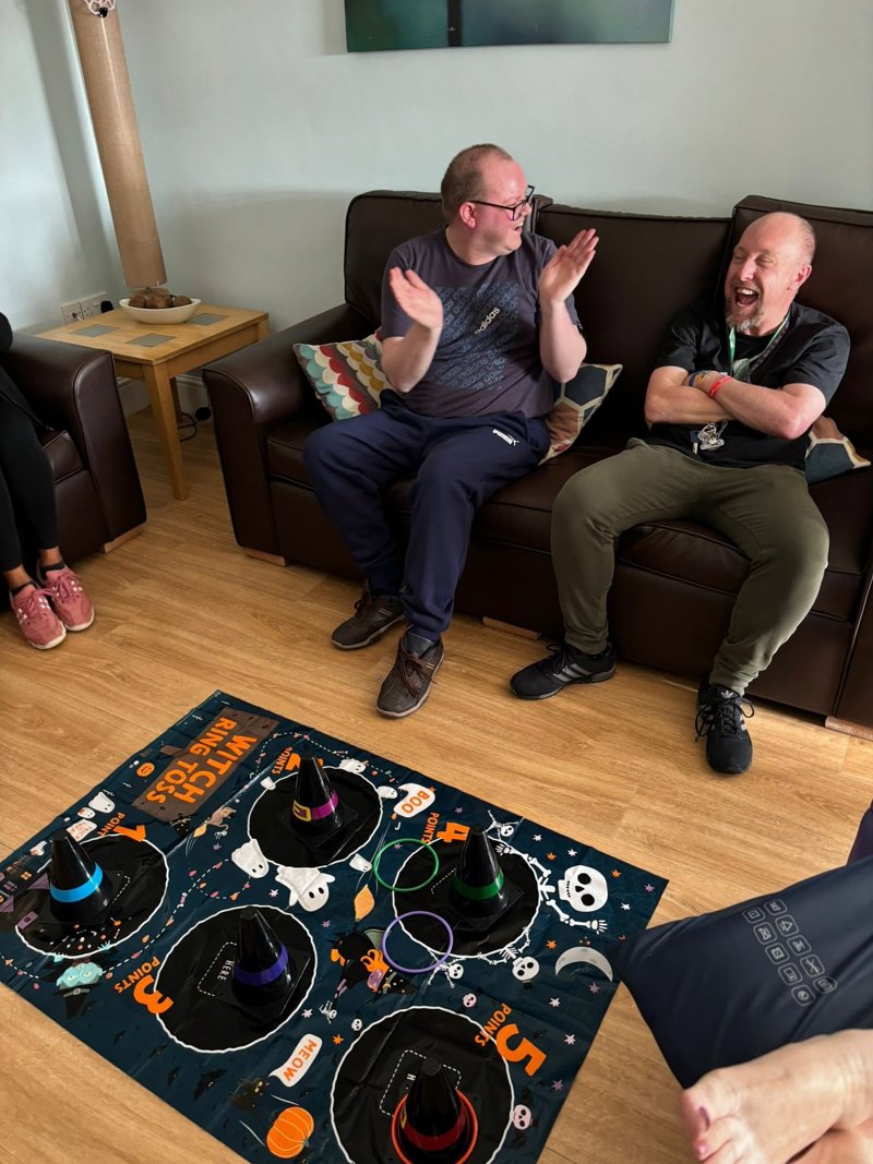 Two men laughing on a sofa beside a Halloween witch ring-toss game in the Oakhill lounge