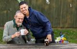 Support worker resting his head on a smiling resident as they share mugs of tea at an outdoor table at Paddocks