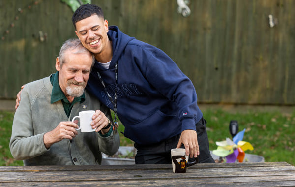 Support worker resting his head on a smiling resident as they share mugs of tea at an outdoor table at Paddocks
